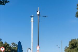 A Synergiewerk smart pole with integrated small cell and LED lighting stands before a drop tower and Ferris wheel in Munich.
