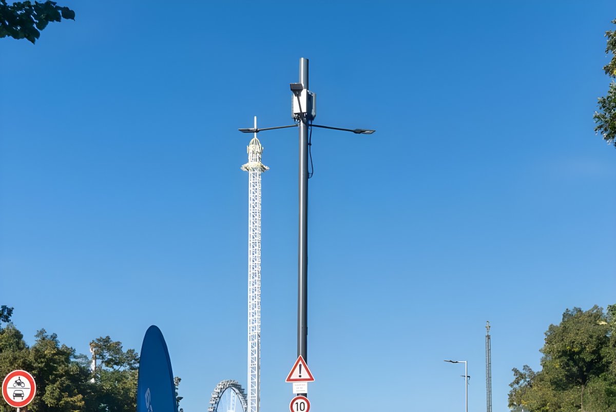 A Synergiewerk smart pole with integrated small cell and LED lighting stands before a drop tower and Ferris wheel in Munich.