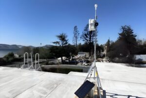Solar-powered air quality monitoring station on a tripod overlooking a residential area with charred trees.