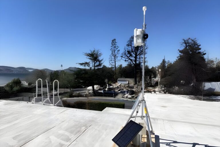Solar-powered air quality monitoring station on a tripod overlooking a residential area with charred trees.