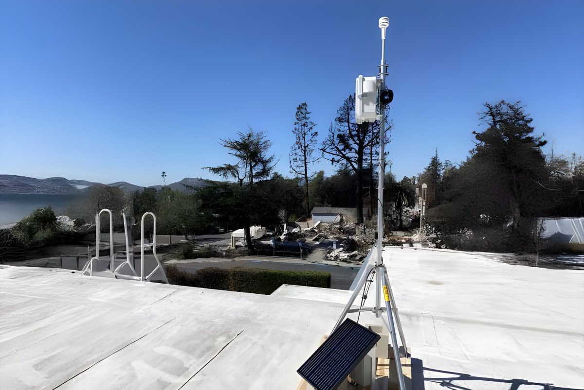 Solar-powered air quality monitoring station on a tripod overlooking a residential area with charred trees.