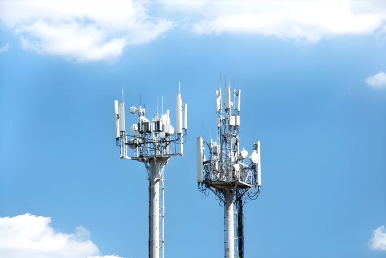 Two metal telecommunications towers with mounted cellular antennas and satellite dishes against a blue sky with clouds.