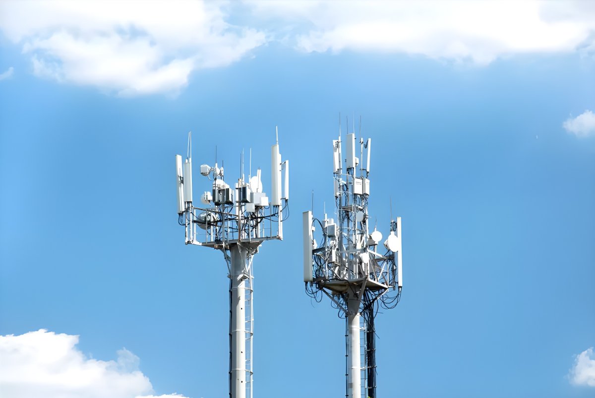 Two metal telecommunications towers with mounted cellular antennas and satellite dishes against a blue sky with clouds.