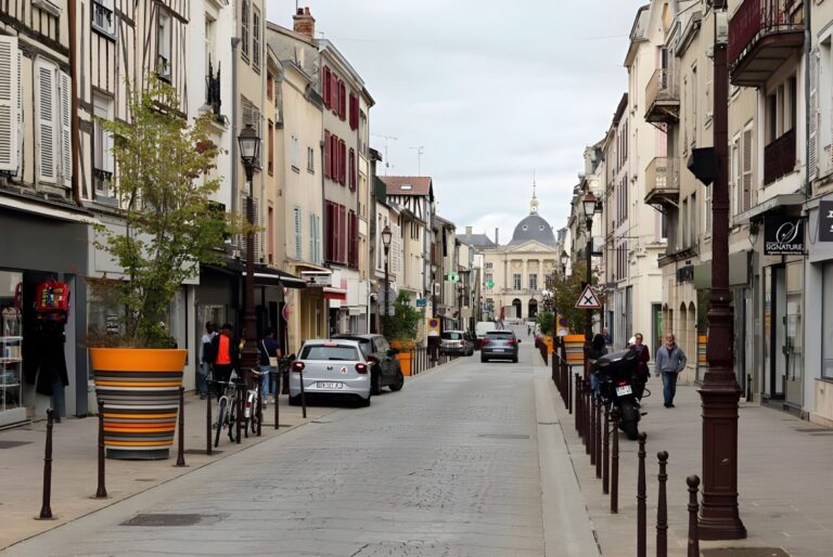 Cobblestone street in Châlons-en-Champagne with parked cars, pedestrians, and historic buildings leading to a dome.