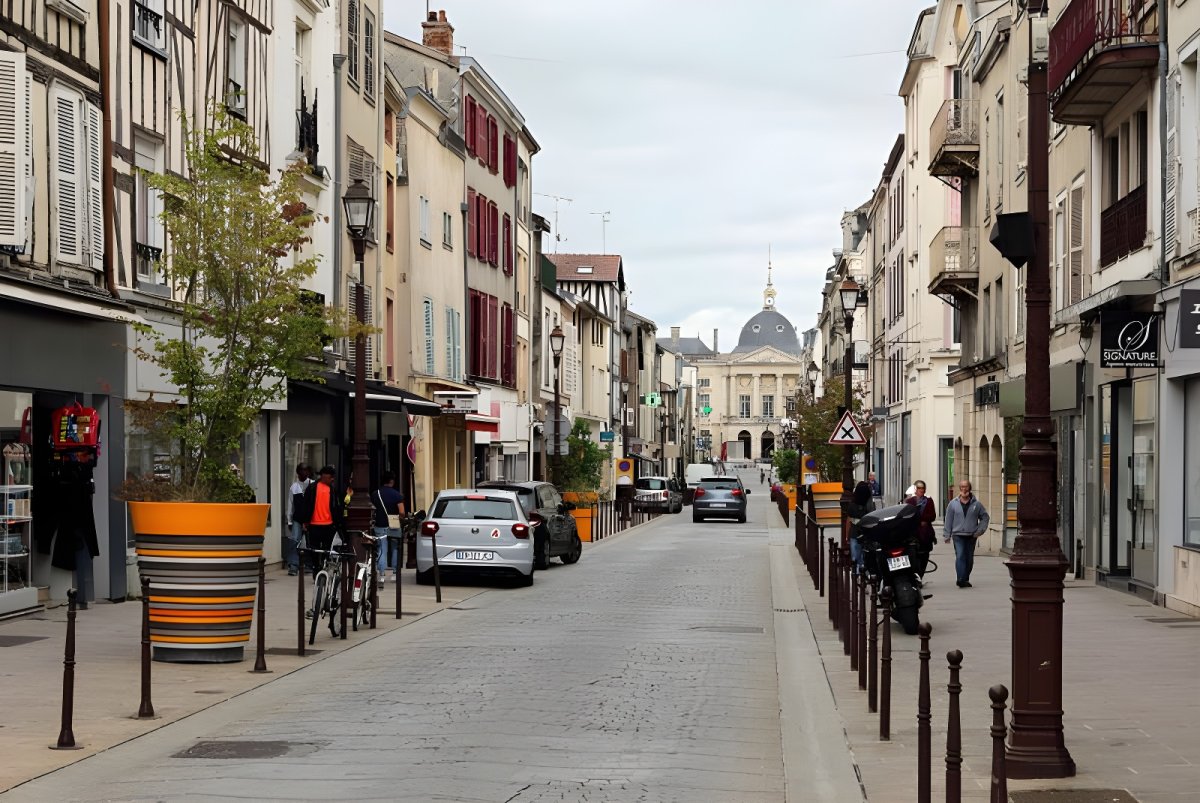 Cobblestone street in Châlons-en-Champagne with parked cars, pedestrians, and historic buildings leading to a dome.