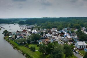 Aerial view of a coastal town with residential houses, a marina, and dense green forests under a cloudy sky.