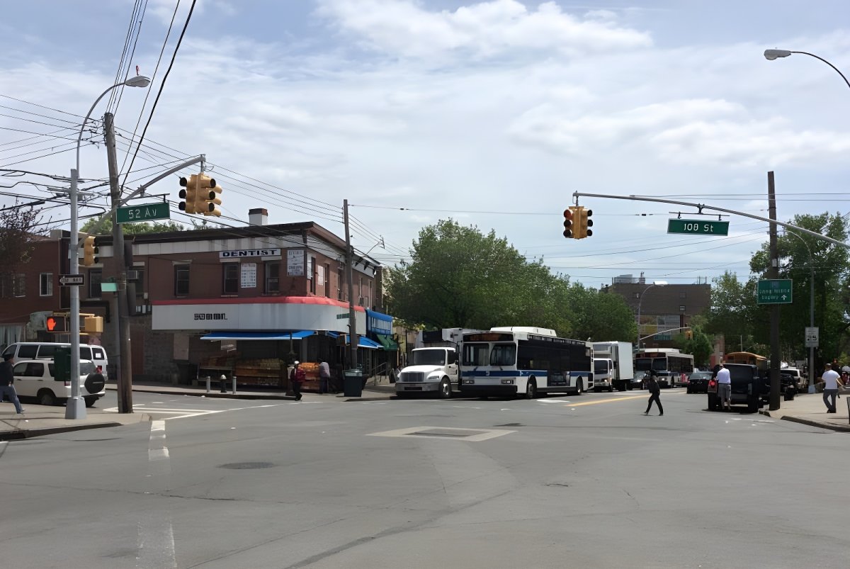 Buses and trucks waiting at a city intersection with overhead traffic lights and street signs under a cloudy sky.