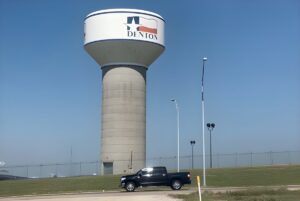 A concrete Denton water tower with a Texas flag logo stands behind a black pickup truck on a clear day.