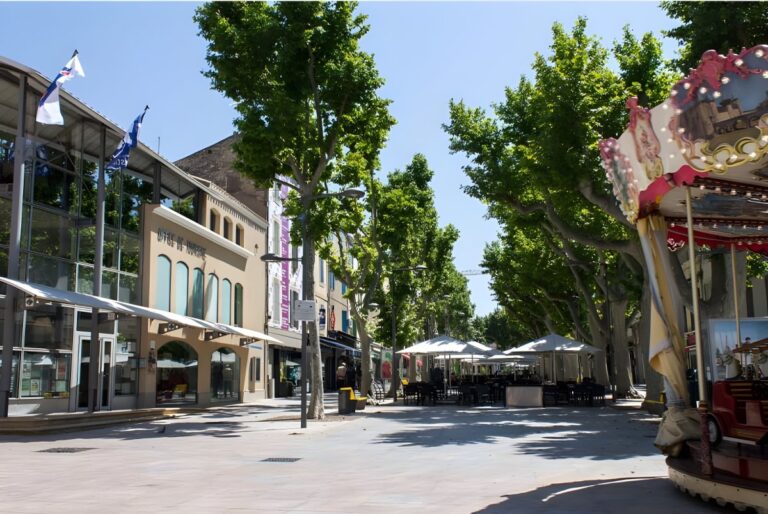 Tree-lined pedestrian plaza in Istres, France, featuring outdoor cafe seating, a carousel, and modern stone buildings.