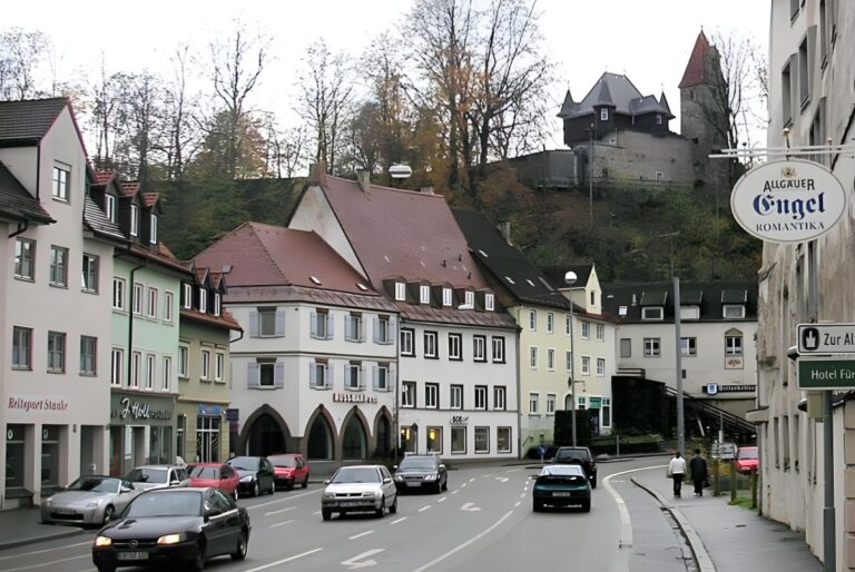 Street view of Kempten, Germany, featuring traditional buildings, parked cars, and Burghalde castle on a wooded hill.