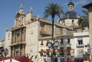 The Church of the Assumption in Llíria, Spain, featuring traditional street lamps and palm trees under a blue sky.