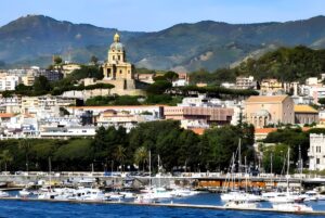 Panoramic view of the Messina harbor and skyline featuring the Sacrario di Cristo Re against a backdrop of green hills.