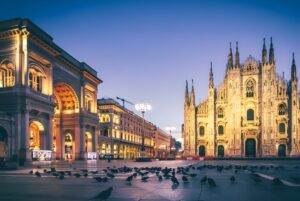 Piazza del Duomo in Milan at dusk featuring the Milan Cathedral, Galleria Vittorio Emanuele II, and street lighting.