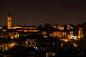 Nighttime view of the Modena skyline featuring illuminated historic buildings, church towers, and warm street lighting.