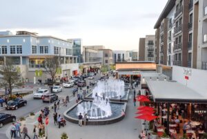 Pedestrians and cars navigate a modern mixed-use development featuring outdoor fountains, storefronts, and apartments.