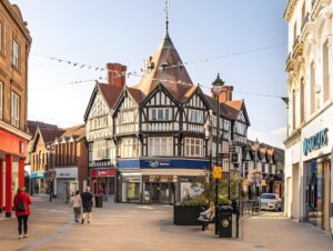 Pedestrians walk through a paved town center in Wrexham featuring half-timbered buildings, a Boots store, and street lamps.