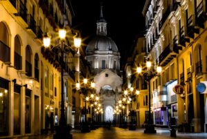 Illuminated street lamps line a narrow Zaragoza street leading toward the domed Basilica of Our Lady of the Pillar at night.