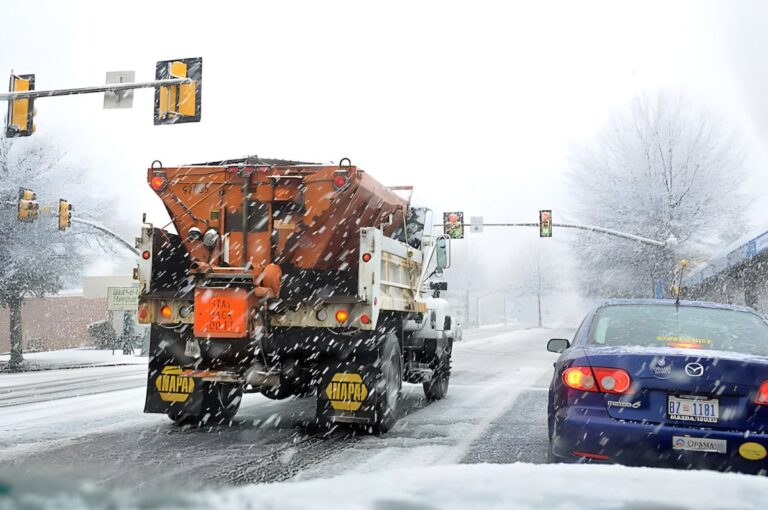 Orange salt spreader truck and a blue car driving through a snowy intersection with traffic lights.
