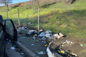 Piles of loose trash and debris scattered along a dirt roadside next to a chain-link fence and grassy embankment.
