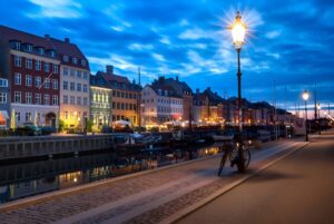Illuminated street lamps line the Nyhavn canal waterfront and colorful townhouses in Copenhagen at dusk.