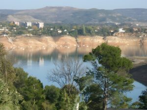 Panoramic view of a reservoir with low water levels and exposed rocky banks near a forested hillside and small town.