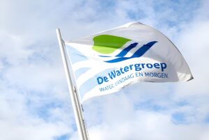 A white De Watergroep flag with a green and blue logo waving on a flagpole against a cloudy blue sky.