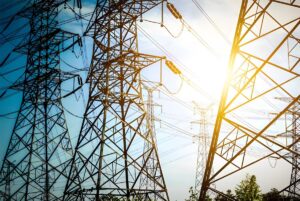 High-voltage steel lattice transmission towers and power lines under a bright sun and blue sky.