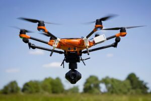Orange and black hexacopter drone with a gimbal camera flying over a green field against a blue sky.