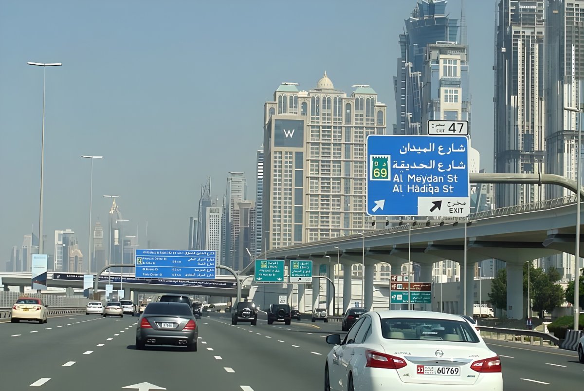 Vehicles traveling on a multi-lane Dubai highway toward skyscrapers with blue Arabic and English overhead road signs.
