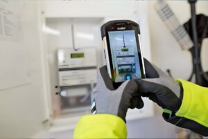 A technician in high-visibility gear and gloves uses a handheld mobile device to photograph a digital smart meter.