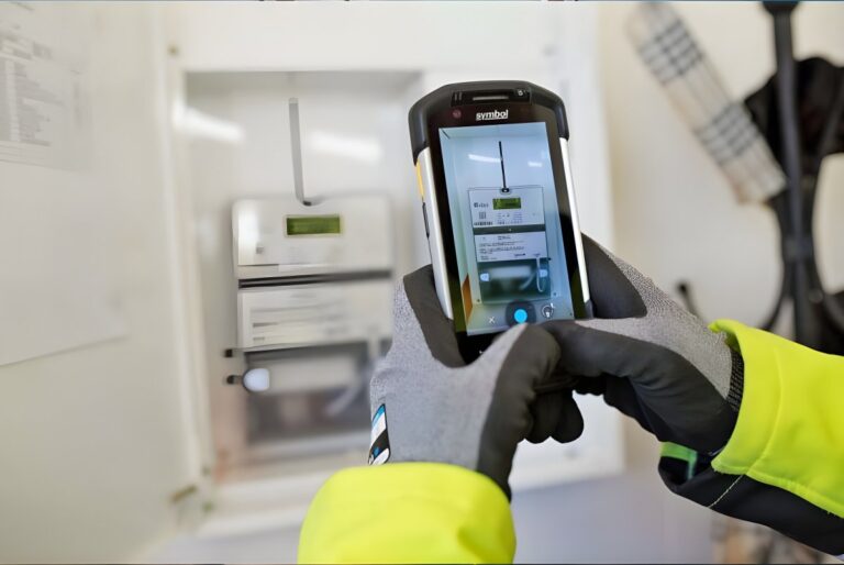 A technician in high-visibility gear and gloves uses a handheld mobile device to photograph a digital smart meter.