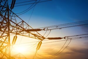 High-voltage steel lattice transmission tower and power lines silhouetted against a bright sunset.