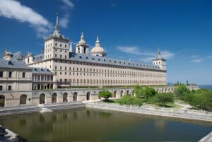 The Royal Seat of San Lorenzo de El Escorial featuring the monastery, stone arches, and reflecting pool under a blue sky.