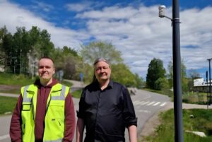 Two men stand on a paved path near a pole mounted with a small white weather sensor under a blue sky.