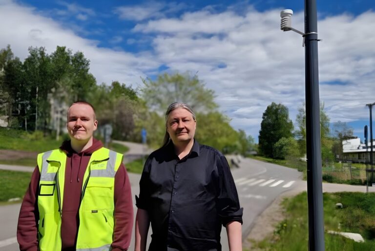 Two men stand on a paved path near a pole mounted with a small white weather sensor under a blue sky.
