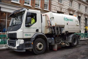 White FM Conway street sweeper truck operating on a torn-up urban road next to a brick building.