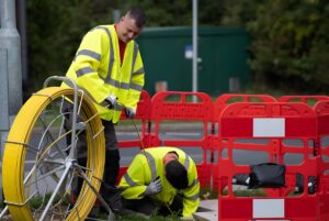 Two technicians in high-visibility jackets install fiber optic cable from a yellow reel into an underground utility manhole.