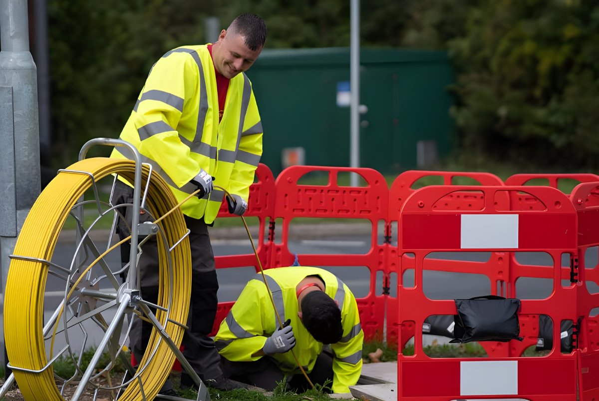 Two technicians in high-visibility jackets install fiber optic cable from a yellow reel into an underground utility manhole.