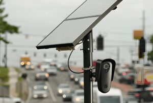 A Flock Safety license plate recognition camera and solar panel mounted on a pole overlooking a busy city road.