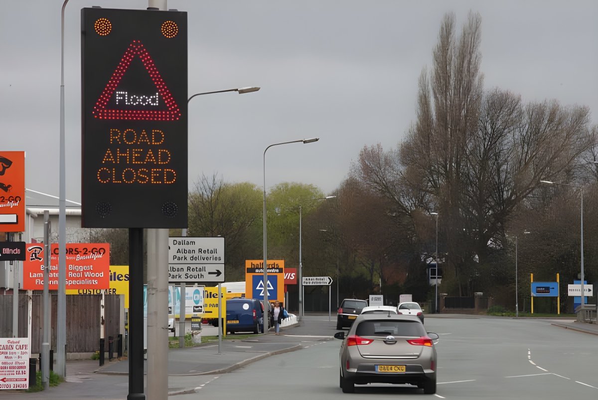 Digital SWARCO road sign displaying a flood warning and road closed message on a street with approaching traffic.