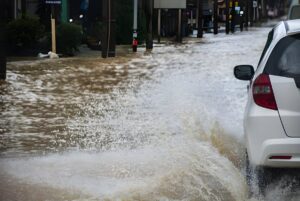 White car driving through a flooded city street with water splashing against the vehicle.