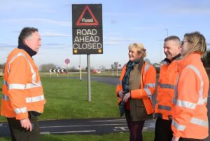 Four people in high-visibility jackets stand near a digital SWARCO road sign displaying a flood warning and road closure.