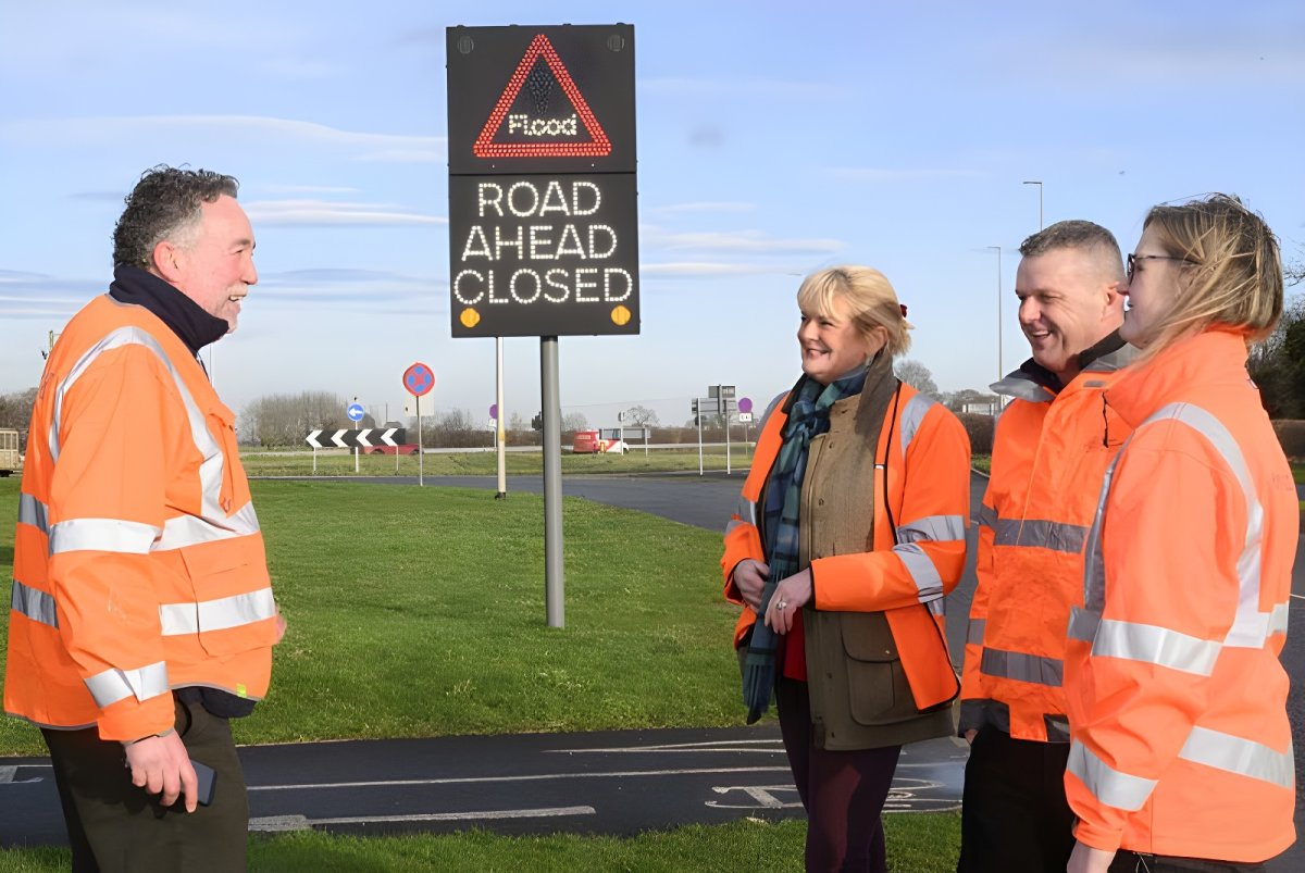 Four people in high-visibility jackets stand near a digital SWARCO road sign displaying a flood warning and road closure.
