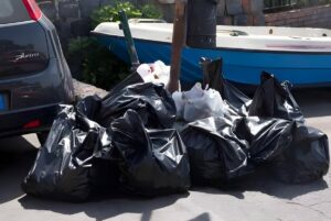 Black plastic trash bags piled on a paved street next to a parked car and a blue boat.