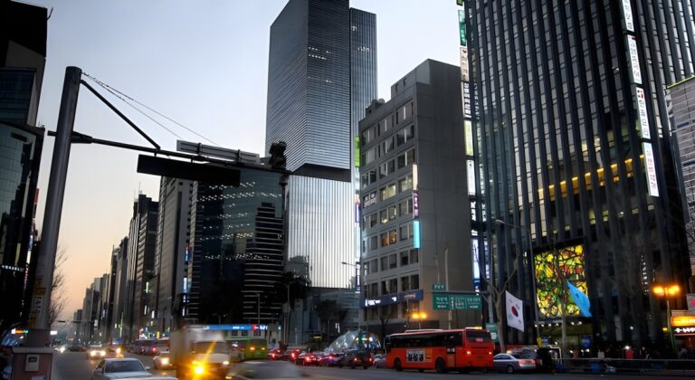 Traffic and buses moving through a dense Seoul city street lined with illuminated skyscrapers at dusk.