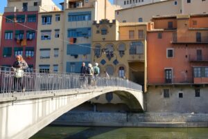 Pedestrians walking across a stone arch bridge over a river toward colorful historic buildings in Girona, Spain.
