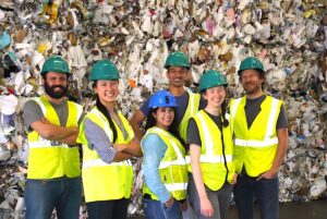 Six workers in hard hats and high-visibility vests stand in front of a large wall of baled recyclable materials.