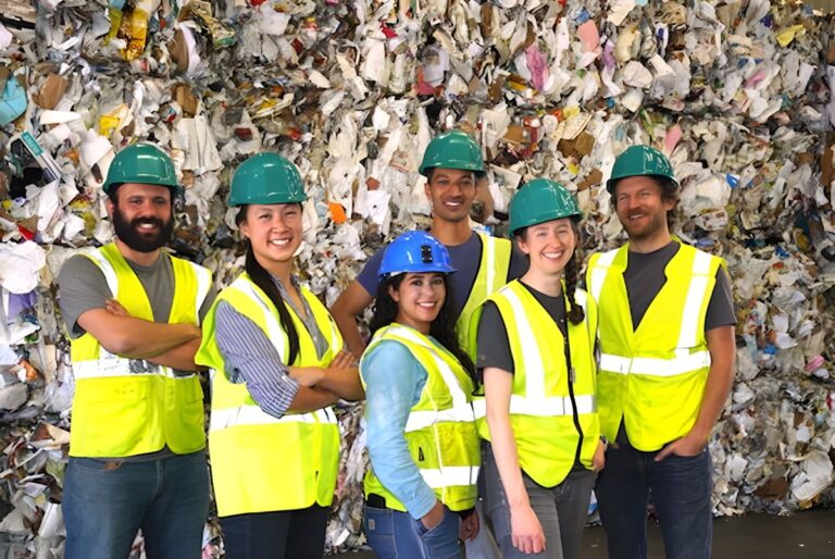 Six workers in hard hats and high-visibility vests stand in front of a large wall of baled recyclable materials.