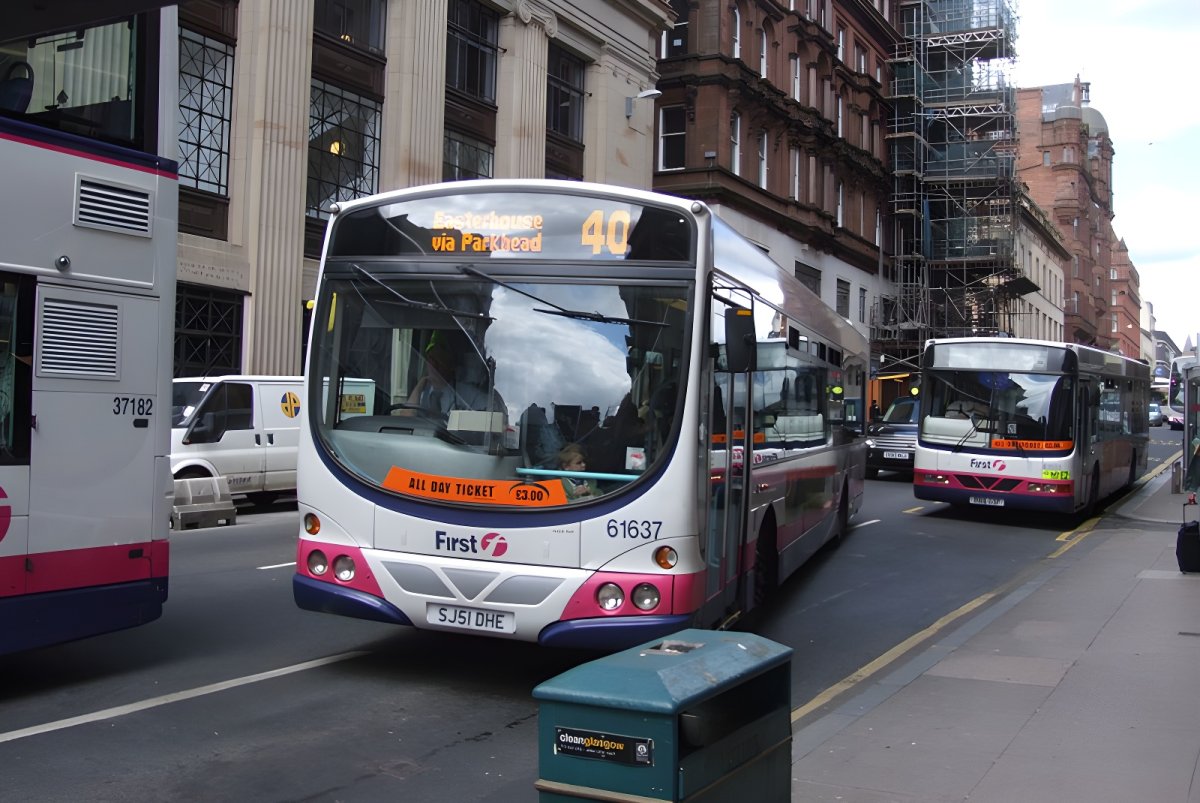 First Bus vehicles driving on a multi-lane city street in Glasgow lined with stone buildings and scaffolding.
