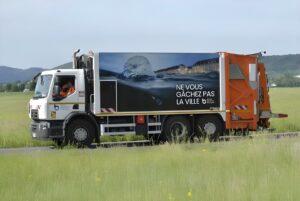 A Grand Besançon Métropole waste collection truck with a Sulo body driving past a grassy field.